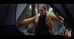 A young man with long red hair and a determined expression crawls out of a tent opening in a misty forest campsite. He wears a yellow parka with a sherpa collar over a black shirt, arms outstretched as he emerges.