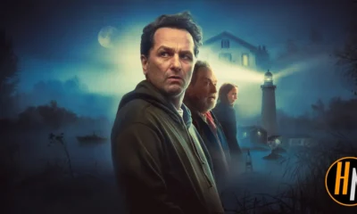 Promotional image for the Apple TV+ series Widow's Bay. A man in a dark hoodie stands in the foreground, looking wary, with an older man and a young woman behind him. A misty coastal scene surrounds them, featuring a lighthouse beam cutting through fog, a house with lit windows, and a small boat on dark water under a full moon. The Horrortainment News logo appears in the lower right corner.