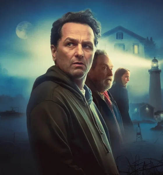 Promotional image for the Apple TV+ series Widow's Bay. A man in a dark hoodie stands in the foreground, looking wary, with an older man and a young woman behind him. A misty coastal scene surrounds them, featuring a lighthouse beam cutting through fog, a house with lit windows, and a small boat on dark water under a full moon. The Horrortainment News logo appears in the lower right corner.