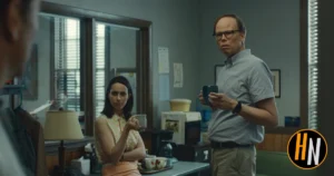 A still from the Apple TV+ series Widow's Bay. In a cluttered, dated office or back room with venetian blinds and wood-paneled walls, a woman with dark hair sits with her arms crossed, holding a small cup and wearing a floral sleeveless top, her expression guarded and skeptical. Beside her stands a tall, thin man in glasses and a gray short-sleeved button-up shirt, holding a coffee mug with a similarly wary look. A third figure is partially visible at the far left edge of the frame. The Horrortainment News logo appears in the lower right corner.