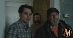 A still from the Apple TV+ series Widow's Bay. Two men stand in the foreground of a dimly lit room with dated floral wallpaper, both staring off-screen with wide, alarmed expressions. The man on the left wears a plaid shirt; the man on the right is older, with a gray beard and a dark cap over an orange shirt. A third man lurks partially visible in the doorway behind them, looking equally unsettled. The Horrortainment News logo appears in the lower right corner.