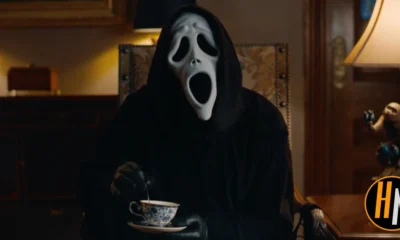 Ghostface from Scary Movie (2026) sits in an ornate, dimly lit parlor room, wearing the iconic white ghost mask and black robe, stirring a delicate blue-and-white china teacup with a black-gloved hand. A vintage lamp and a small decorative figurine are visible in the background. The Horrortainment News logo appears in the lower right corner.