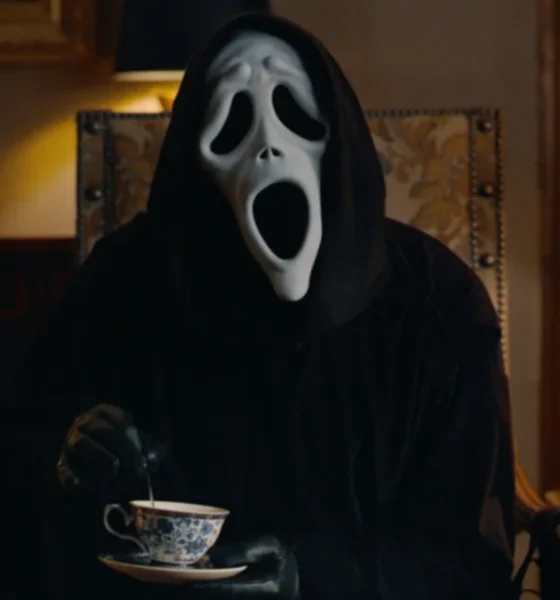 Ghostface from Scary Movie (2026) sits in an ornate, dimly lit parlor room, wearing the iconic white ghost mask and black robe, stirring a delicate blue-and-white china teacup with a black-gloved hand. A vintage lamp and a small decorative figurine are visible in the background. The Horrortainment News logo appears in the lower right corner.