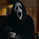 Ghostface from Scary Movie (2026) sits in an ornate, dimly lit parlor room, wearing the iconic white ghost mask and black robe, stirring a delicate blue-and-white china teacup with a black-gloved hand. A vintage lamp and a small decorative figurine are visible in the background. The Horrortainment News logo appears in the lower right corner.