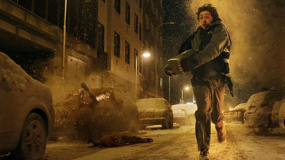 A young man runs toward the camera down a snow-covered street at night, clutching a green case in both arms with a submachine gun at his hip. Dirt and debris explode behind him beside a wrecked car, and a body lies in the snow.