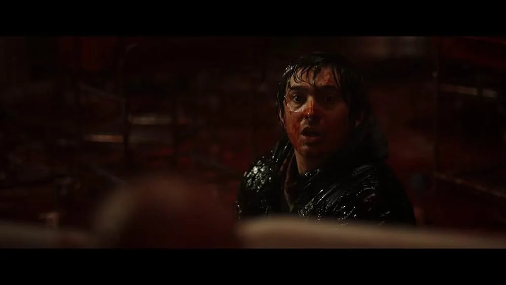 Close-up of a wide-eyed man with wet dark hair, his face and black jacket smeared with blood, in a dim, red-tinted room with metal racks in the background.