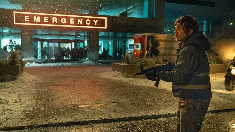 A man in a dark jacket with reflective stripes holds a submachine gun in a snowy forecourt outside a hospital. A glowing red "EMERGENCY" sign hangs overhead, an ambulance is parked nearby, and a crowd of silhouetted people gathers at the entrance.
