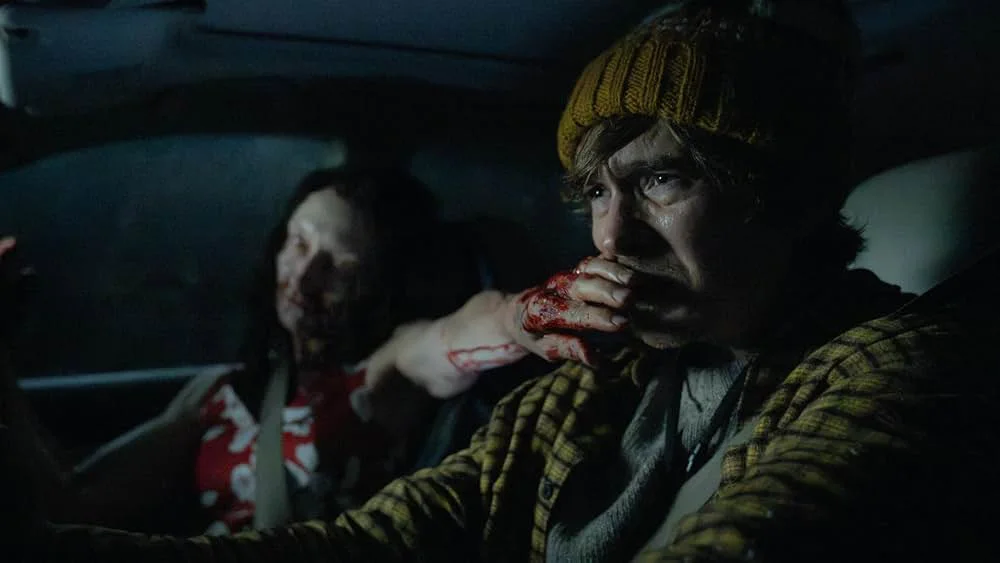 Inside a dark car at night, a man in a mustard knit beanie and yellow plaid jacket, looking distressed. Beside him, a pale woman in a red floral dress with blood on her face reaches toward him, her seatbelt still on, she presses a blood-covered hand over his mouth.
