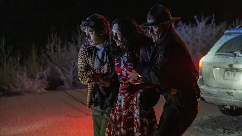 On a snowy roadside at night, lit by a red glow, a young man in a plaid jacket and a dark-haired woman in a red floral dress are helped along by a sheriff in a campaign hat. A snow-dusted white station wagon sits behind them.