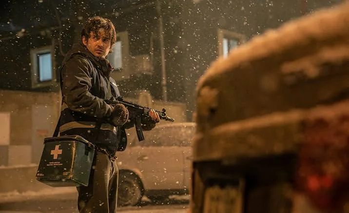 A man in a dark winter jacket with reflective stripes stands on a snowy night street with a submachine gun in one hand and a green first-aid case with a white cross in the other. Snow falls around him, and a snow-covered vehicle is blurred in the foreground.