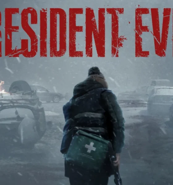 Promotional artwork with the "RESIDENT EVIL" title in distressed red lettering across the top. Below it, a figure seen from behind, wearing a beanie, a dark winter jacket with reflective stripes, and a green first-aid case slung at their side, stands in a heavy snowstorm facing a road littered with wrecked cars. One car lies overturned and burning, and flames burn at the right edge of the frame. A small orange "HN" logo sits in the bottom-right corner.